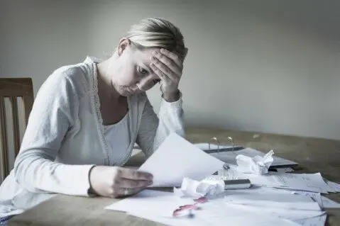 Financial stress from foreclosure process Stressed homeowner sitting at a table, holding her head in frustration while reviewing foreclosure notice of default documents, surrounded by scattered papers, highlighting the emotional toll of the foreclosure process