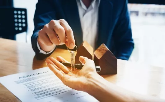 Putting an Inherited Property Under your name Picture of a lawyer in a blue suit, sitting accross the table from a heir inheriting a property. Picture shows lawyer handing off the key over to the heir after putting the property under his name and documents splayed out on the table.