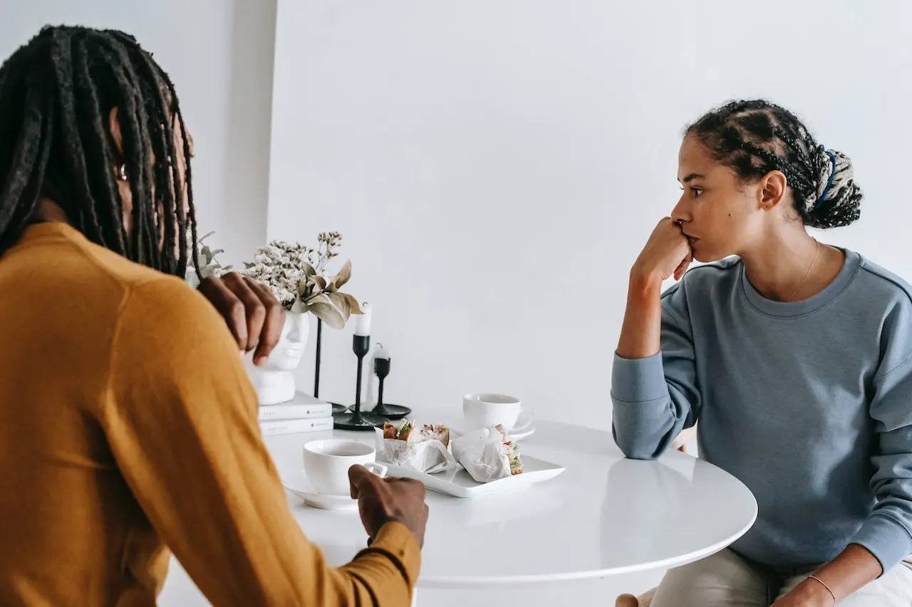 Siblings Argue over inherited house : Two siblings sit at a white table, engaged in a tense conversation about what to do with their inherited house. One, wearing a mustard-colored sweater, gestures while speaking, while the other, in a blue sweatshirt, rests their chin on their hand, looking frustrated and deep in thought. The setting is minimalistic, with coffee cups and sandwiches on the table, and a vase of dried flowers in the background.