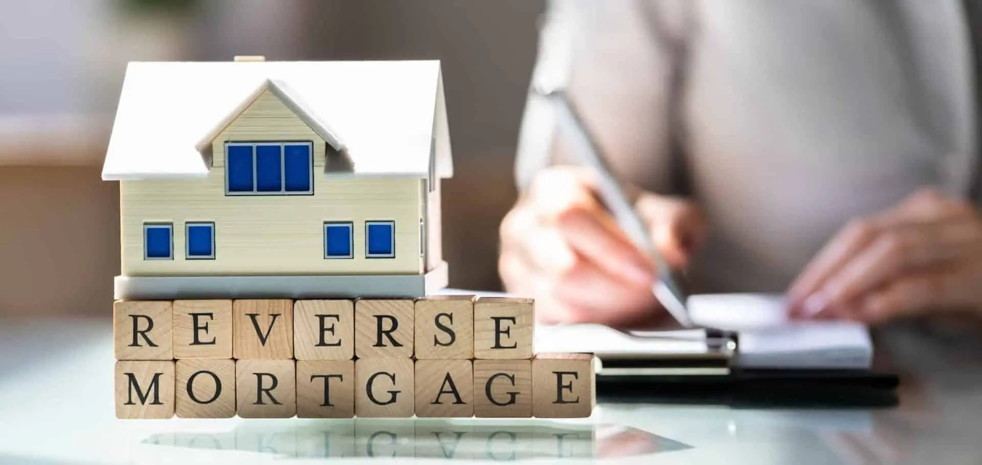 Model house sitting on wooden blocks spelling “reverse mortgage” with a person signing paperwork in the background, symbolizing the process of selling a home with a reverse mortgage.
