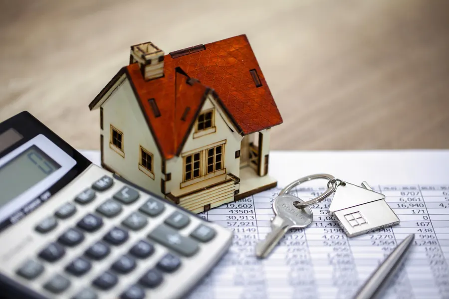 Photo of a calculator and a model home sitting on top of a printed out copy of a spreadsheet, laid on top of a desk, highlighting how wrap around mortgages work in a real estate transaction